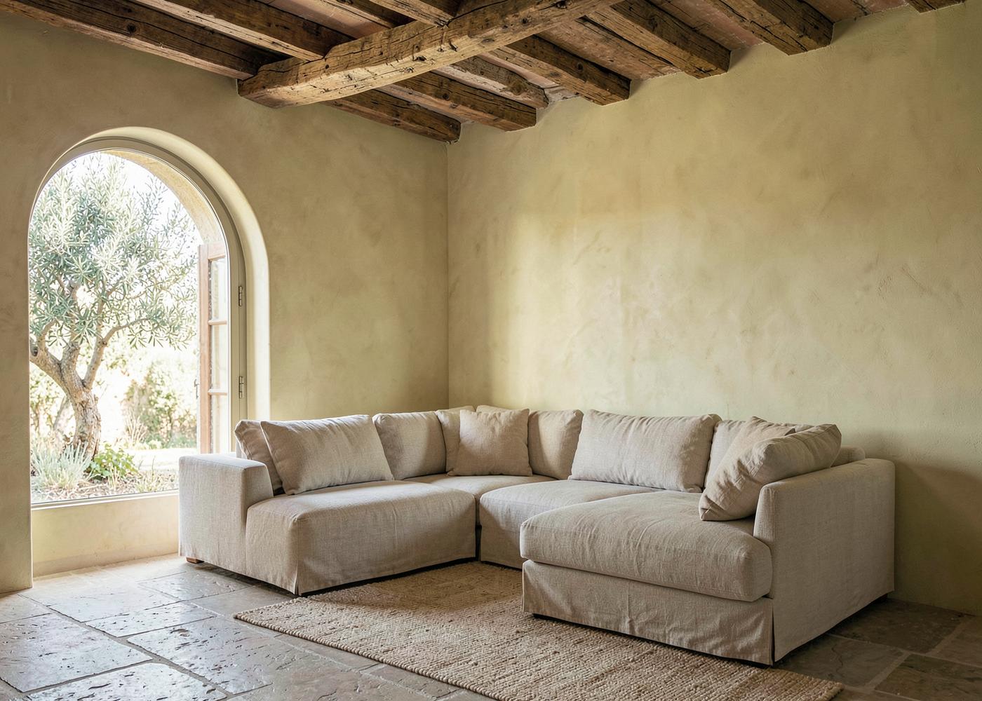 Lime plaster living room with oak beams, low linen sofa and arched window onto an olive tree in a Luberon mas furnished by Maison Quari Vera